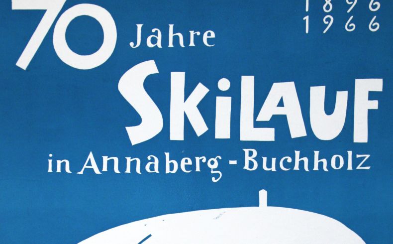 Plakat mit einem angedeuteten weißen Berg, links rote und blaue Fahnen und eine geschwungene Linie von oben nach unten, rechts zwei rote Ski und ein blauer Skistock, oben in einer blauen Fläche die Schrift 70 Jahre Skilauf in Annaberg-Buchholz 1896 - 1966