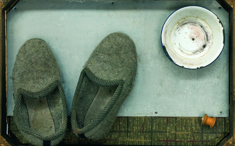 Materialbild, in einer hölzernen (Obst-)kiste sind auf einem Blech links zwei Filzhausschuhe arrangiert, rechts oben eine alte emaillierte Schüssel mit blauem Rand, drunter eine leere Garnspule